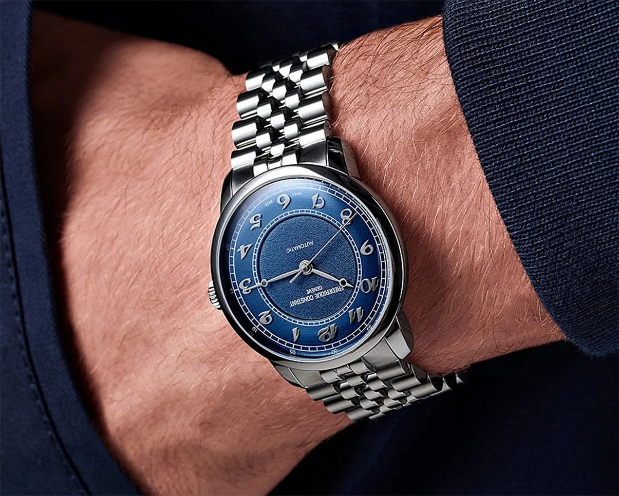 Silver wristwatch with a blue dial featuring stylized Arabic numerals and a textured center, worn on a wrist with a silver metal link bracelet. The watch face displays the brand name "Frederique Constant Genève" and the word "Automatic." The wearer is dressed in a dark blue long-sleeve garment.
