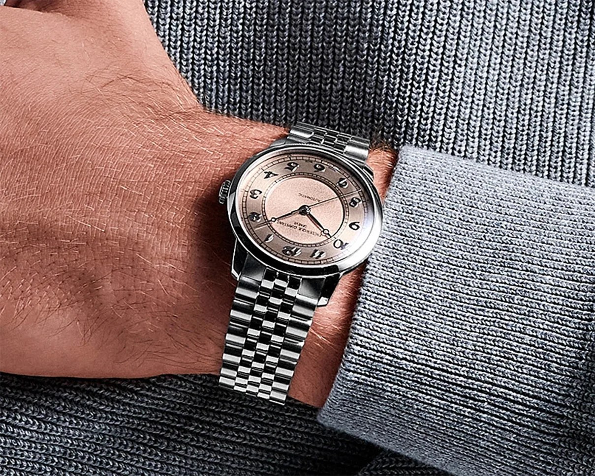 Silver wristwatch with a round, light pink dial featuring black Arabic numerals and a stainless steel link bracelet, worn on a wrist with a gray knitted sweater.