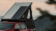 A red pickup truck with a rooftop tent mounted on top. The tent is partially open, featuring a triangular shape with a ladder attached to the side for access. The background shows a blurred natural landscape with hills and a soft, muted sky.
