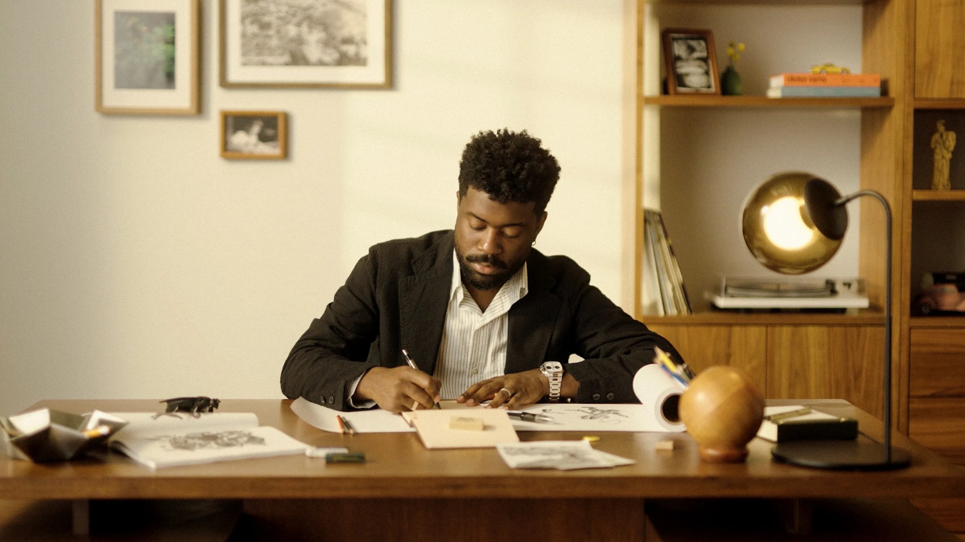 A man with curly hair and a beard, wearing a black blazer and a white striped shirt, is sitting at a wooden desk drawing on paper. The desk has various items including papers, pencils, a wooden spherical object, and a modern desk lamp with a round golden shade. Behind him is a wooden shelving unit with books, framed photos, and decorative items. The wall behind has three framed pictures.