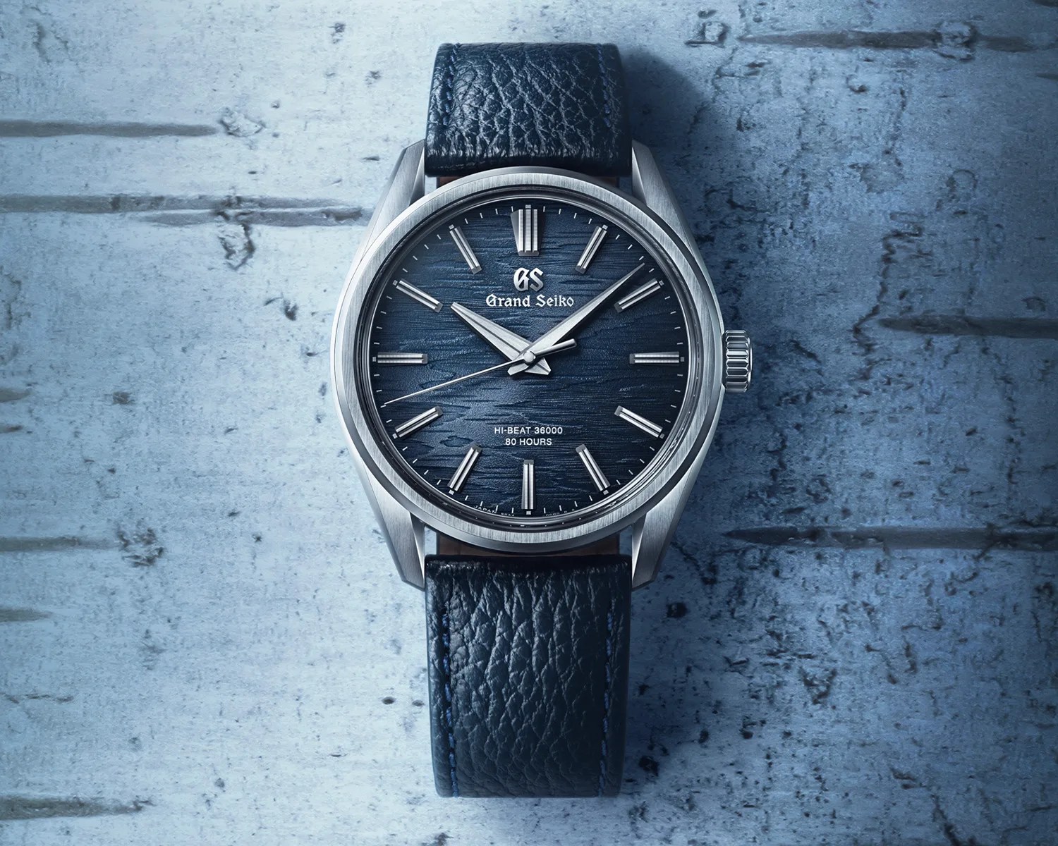 Grand Seiko wristwatch with a textured dark blue dial, silver hour markers, and silver hands. The watch has a stainless steel case and a black leather strap with visible grain. The dial features the Grand Seiko logo at the top and text indicating "HI-BEAT 36000" and "80 HOURS" near the bottom. The background is a textured blue surface.