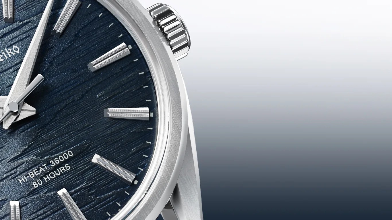 Close-up of a Grand Seiko watch face with a textured dark blue dial, silver hour markers, and hands. The text "HI-BEAT 36000 80 HOURS" is visible on the dial. The watch case and crown are made of polished silver metal.