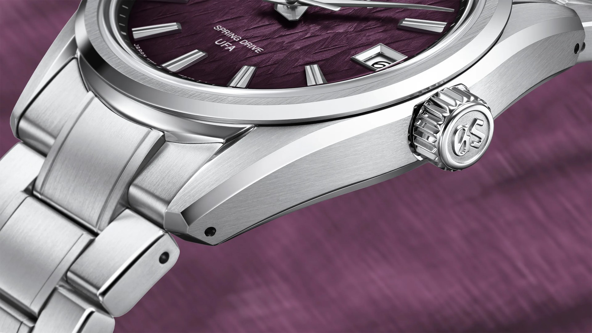Close-up of a luxury stainless steel wristwatch with a textured purple dial. The watch features silver hour markers, a date window at the 3 o'clock position, and a crown engraved with the initials "GS." The dial includes the text "SPRING DRIVE" and "UFA." The watch bracelet is also stainless steel with a brushed finish.