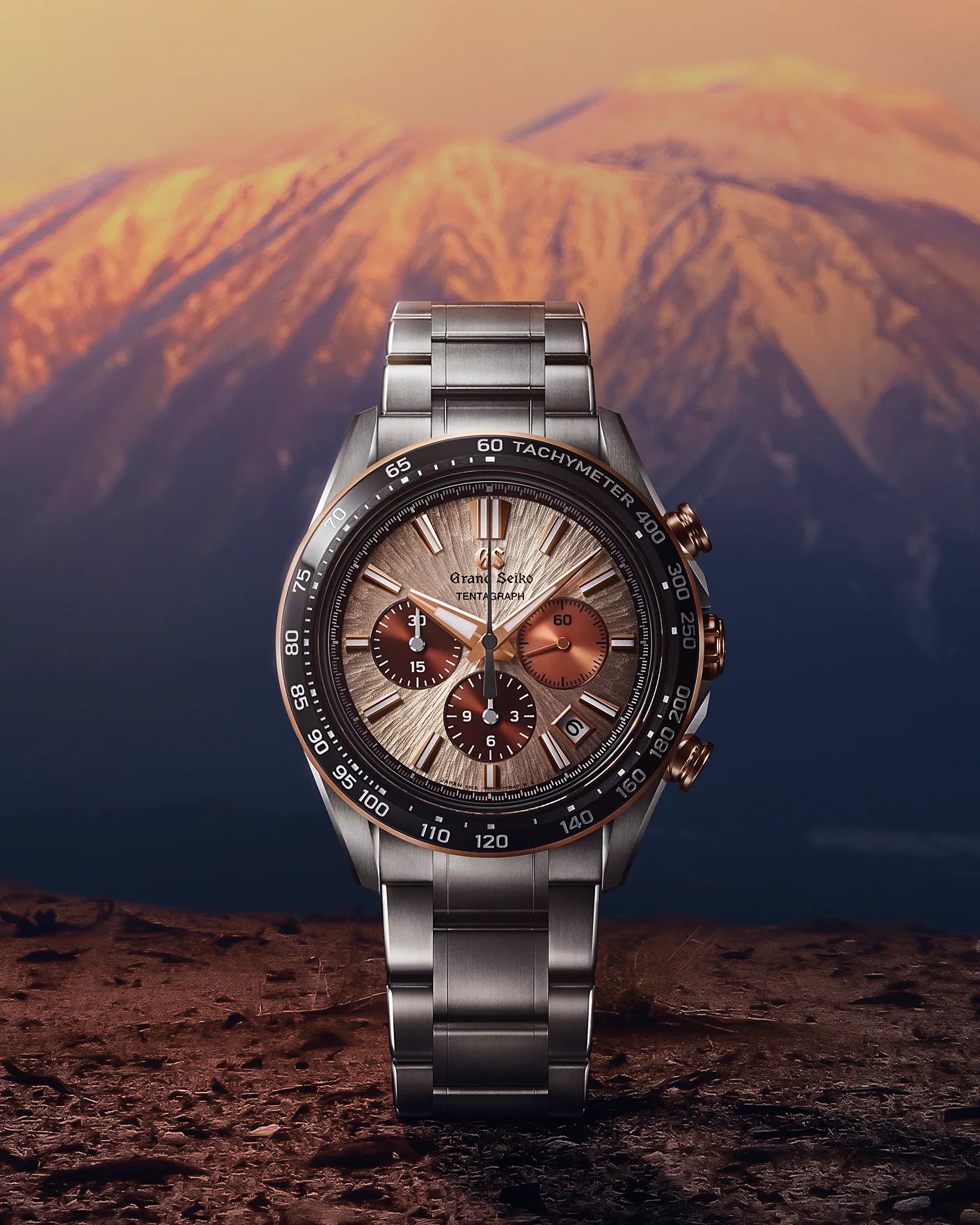 Grand Seiko chronograph watch with a stainless steel bracelet, a textured bronze dial featuring three subdials, gold-tone hands and hour markers, a date window at 4 o'clock, and a black tachymeter bezel. The background shows a softly lit mountain landscape at sunset.