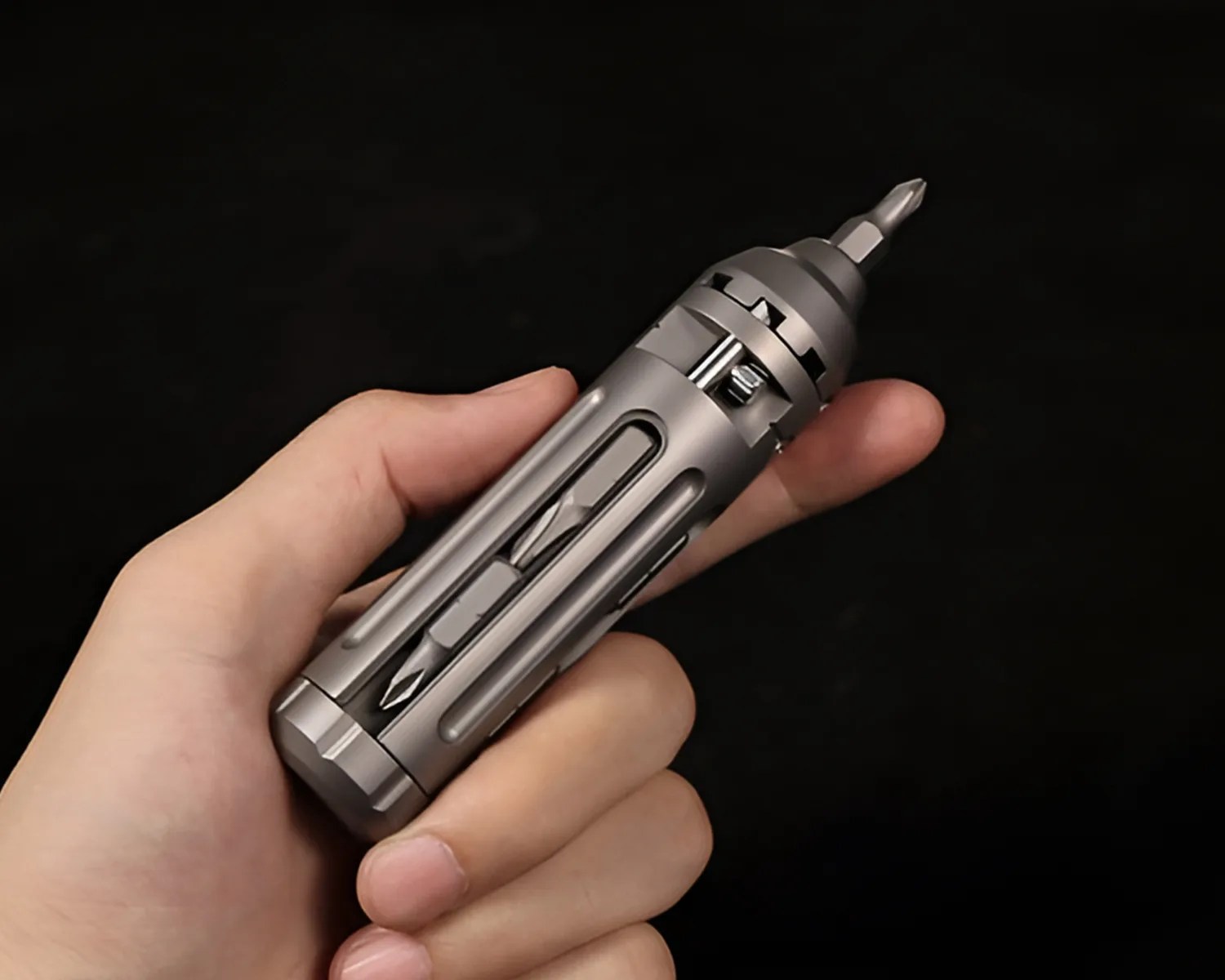 A compact, metallic multi-bit screwdriver held in a hand, featuring a cylindrical design with multiple screwdriver bits stored inside its body. The tool has a sleek, silver-gray finish and a bit attached at the top.