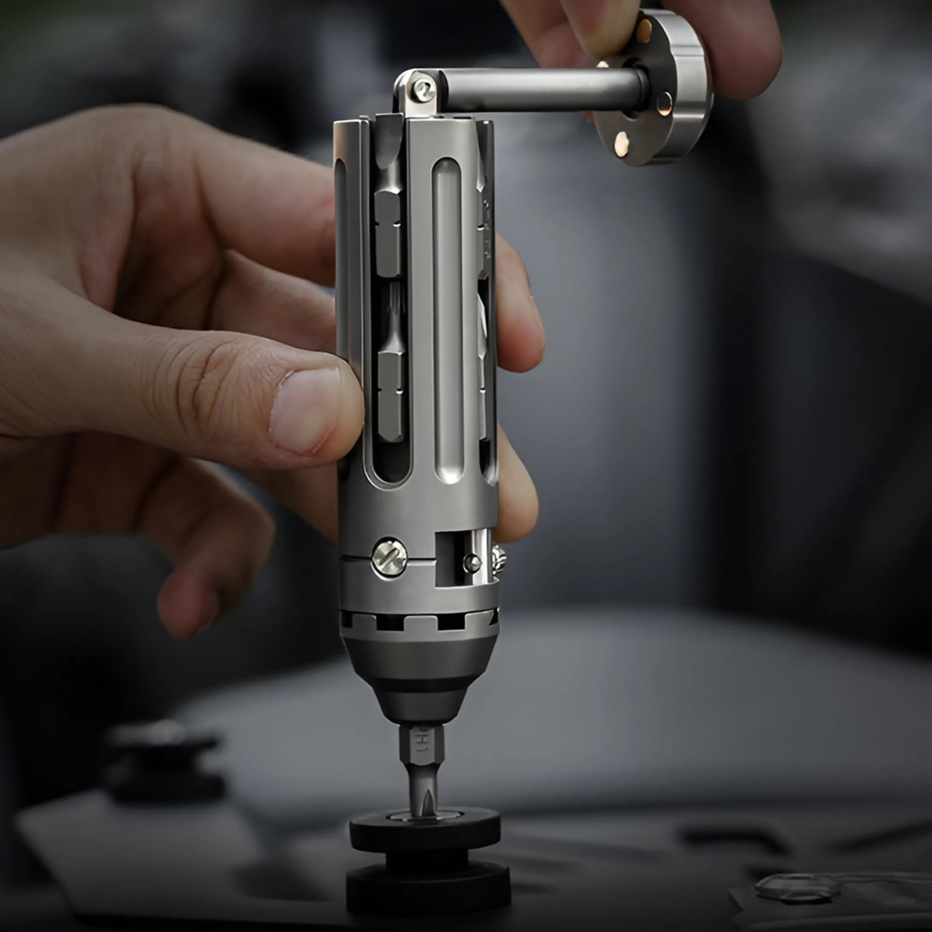 A hand is holding a compact, cylindrical multi-tool with a metallic finish. The tool has various bits stored inside its body and a detachable handle being inserted or removed from the top. The tool is positioned vertically with a screwdriver bit engaged at the bottom, resting on a black surface.