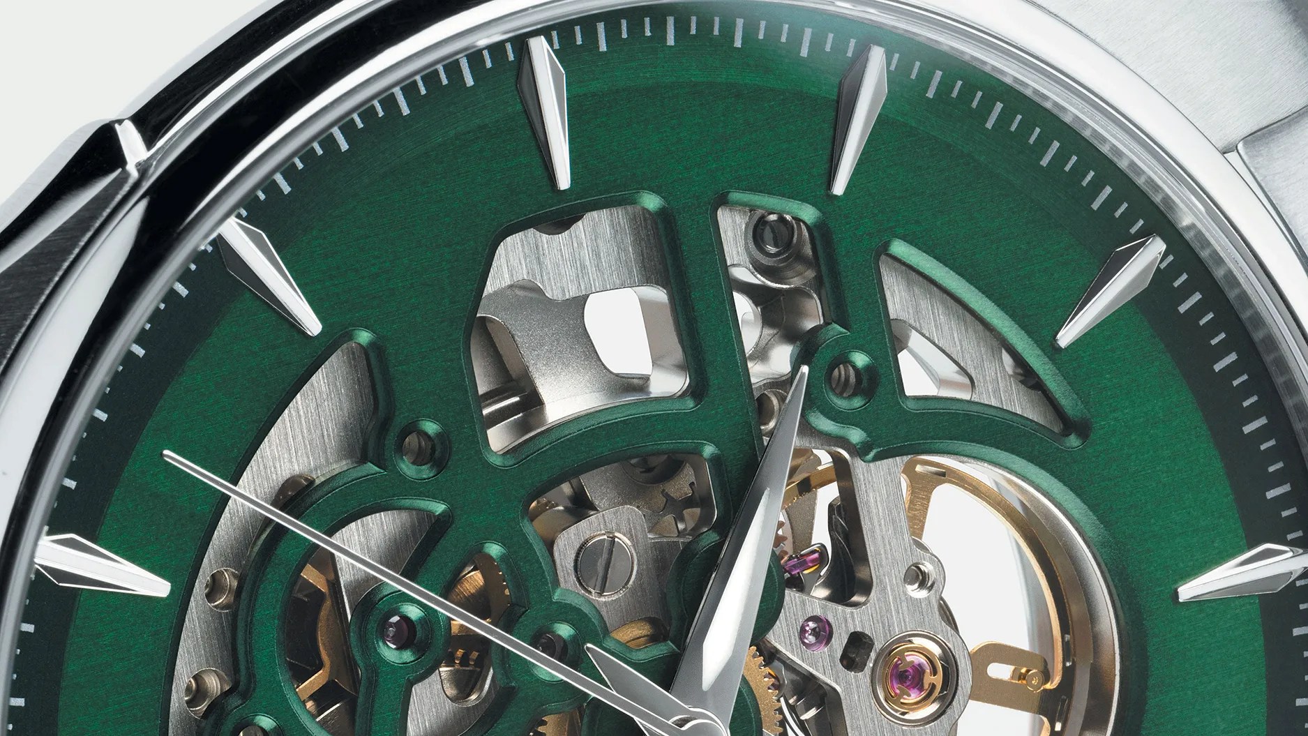 Close-up of a mechanical watch face with a green outer ring and silver hour markers. The watch has visible internal gears and components through cut-out sections in the dial. The hour and minute hands are silver, and there is a thin silver second hand. The watch casing is metallic with a polished finish.