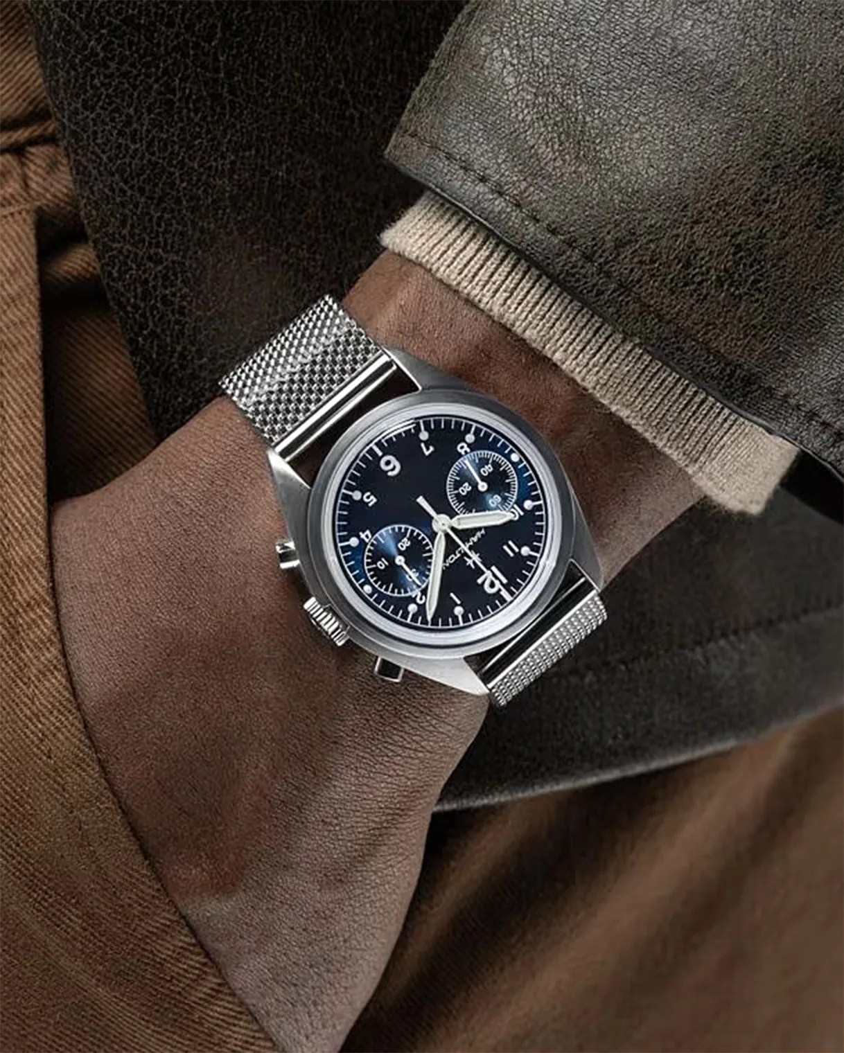 Steel wristwatch with a black dial featuring white numerals and two subdials, worn on a wrist with a silver mesh band. The person is dressed in a brown jacket and beige sweater.