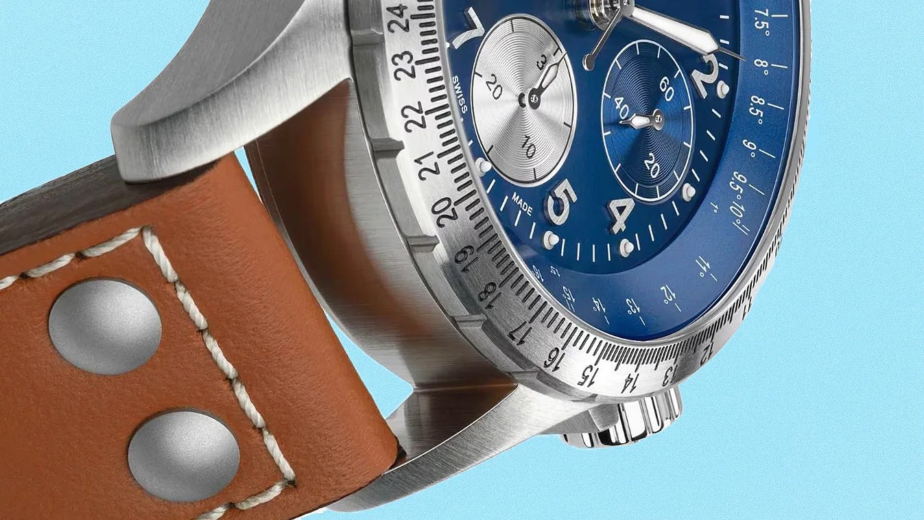 Close-up of a wristwatch featuring a blue dial with two subdials, silver hour markers, and white hands. The watch has a brushed stainless steel case with a rotating bezel marked with numbers from 13 to 24. It is paired with a brown leather strap secured by two metal rivets and white stitching. The background is a solid light blue color.