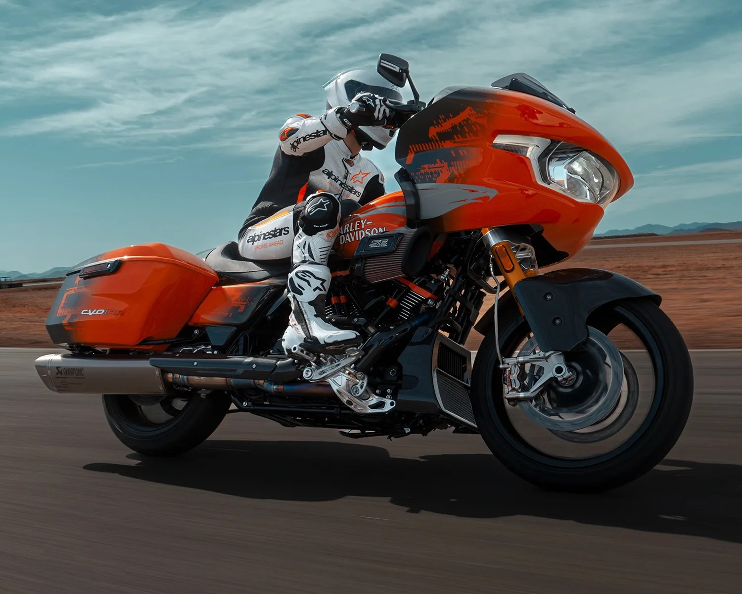 A rider in white and black Alpinestars racing gear is riding an orange Harley-Davidson motorcycle with black and gray accents on a paved road. The motorcycle features a large front fairing, saddlebags, and a visible Akrapovič exhaust system. The background shows a clear sky with some clouds and a barren, desert-like landscape.