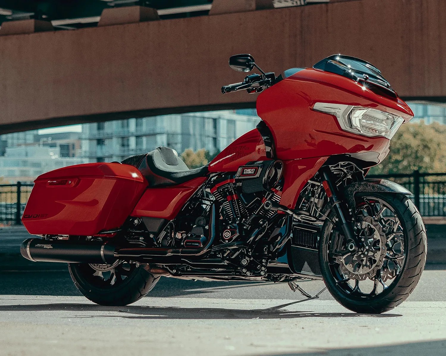 Red touring motorcycle with black accents, featuring a large front fairing, saddlebags, and a comfortable seat. The bike has a prominent engine with "121" displayed on it and is parked on a concrete surface under an overpass with buildings and trees in the background.