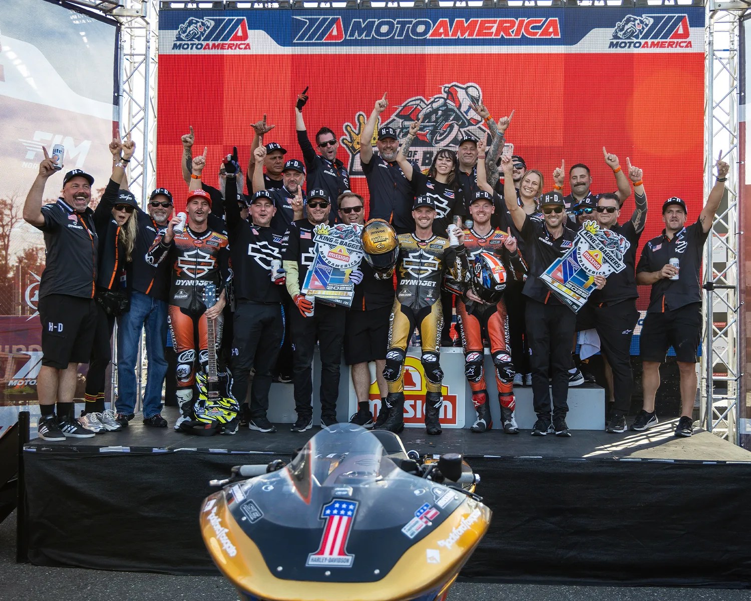 A large group of people, including motorcycle racers in racing suits and team members in black shirts and caps, are celebrating on a podium with a red "MOTOAMERICA" backdrop. Two racers in the front hold helmets and trophies, while others raise their index fingers in a number one gesture. A racing motorcycle with a number one decal is visible in the foreground.