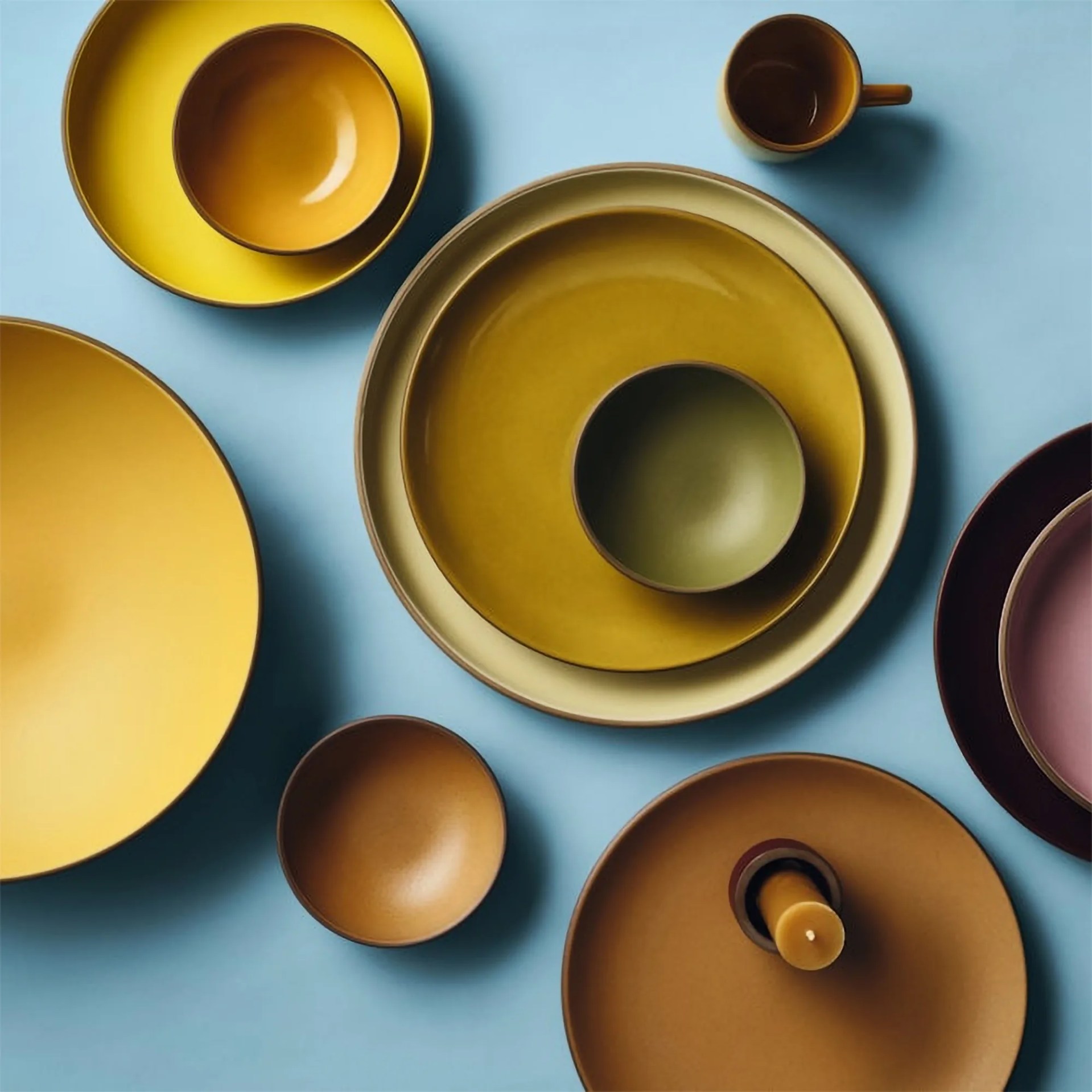 A collection of ceramic tableware arranged on a light blue surface, including various bowls, plates, and a mug in shades of yellow, mustard, olive green, brown, and mauve. One brown plate holds a mustard-colored candle. The items are nested and spaced with a minimalist aesthetic.