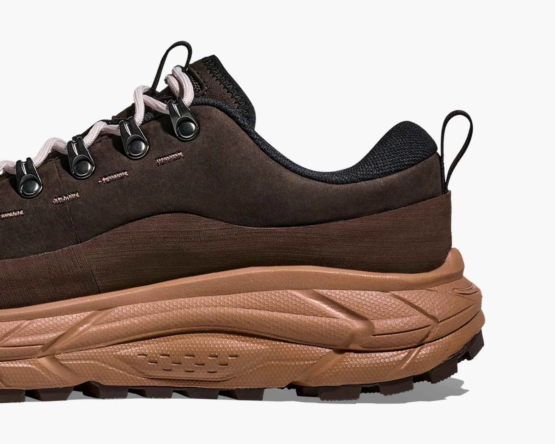Close-up of the back half of a brown hiking shoe with a thick, textured tan sole. The upper part is dark brown with black mesh around the ankle and black metal eyelets for the light pink laces. A black pull tab is attached at the heel.