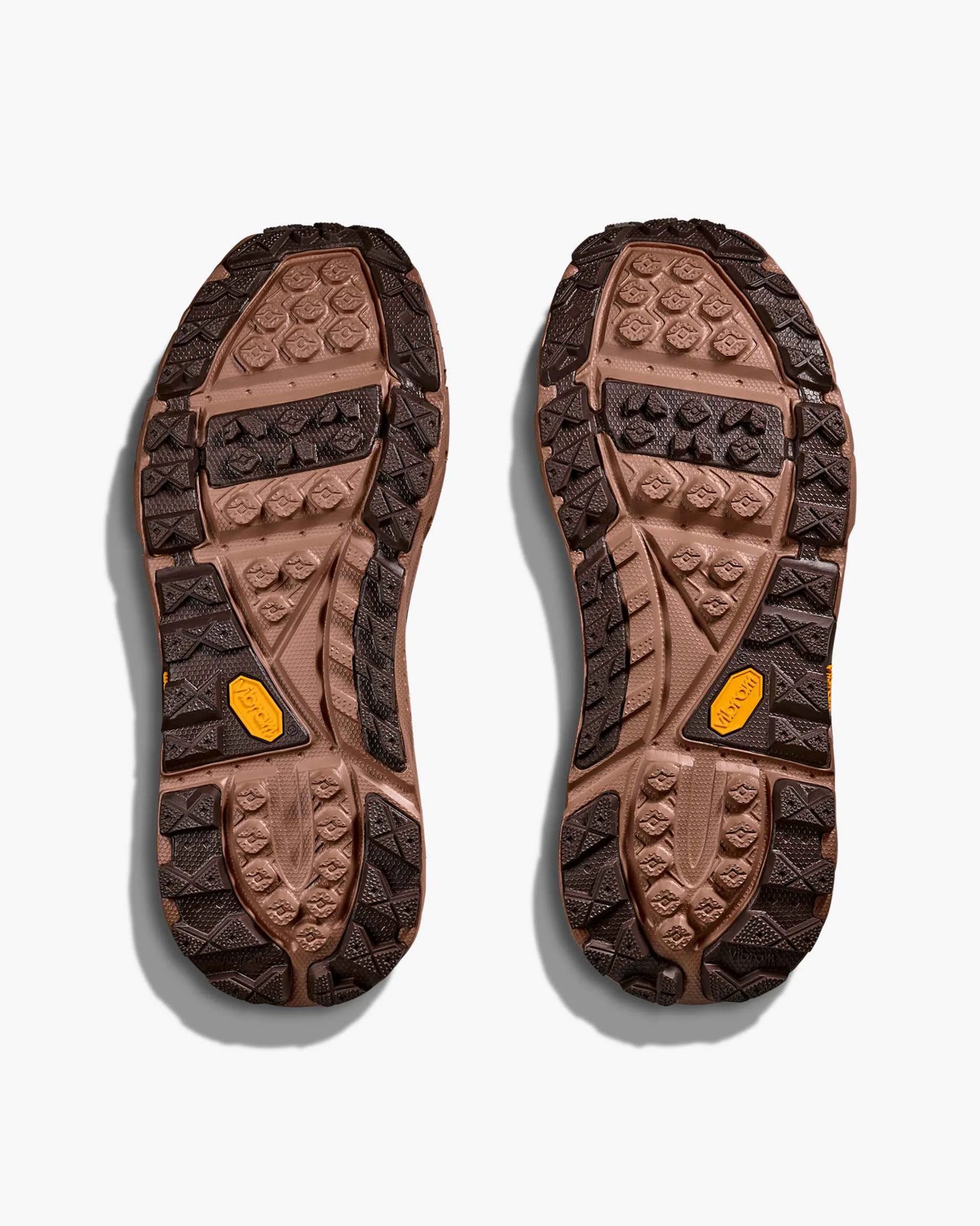 Outsole of a pair of shoes shown from the bottom, featuring a rugged tread pattern in brown and dark brown colors with a yellow Vibram logo on each sole. The design includes various textured sections and lugs for traction.