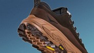 Brown and black hiking shoe with a rugged Vibram sole, featuring metal eyelets for laces and a pull tab at the heel, set against a blue gradient background.
