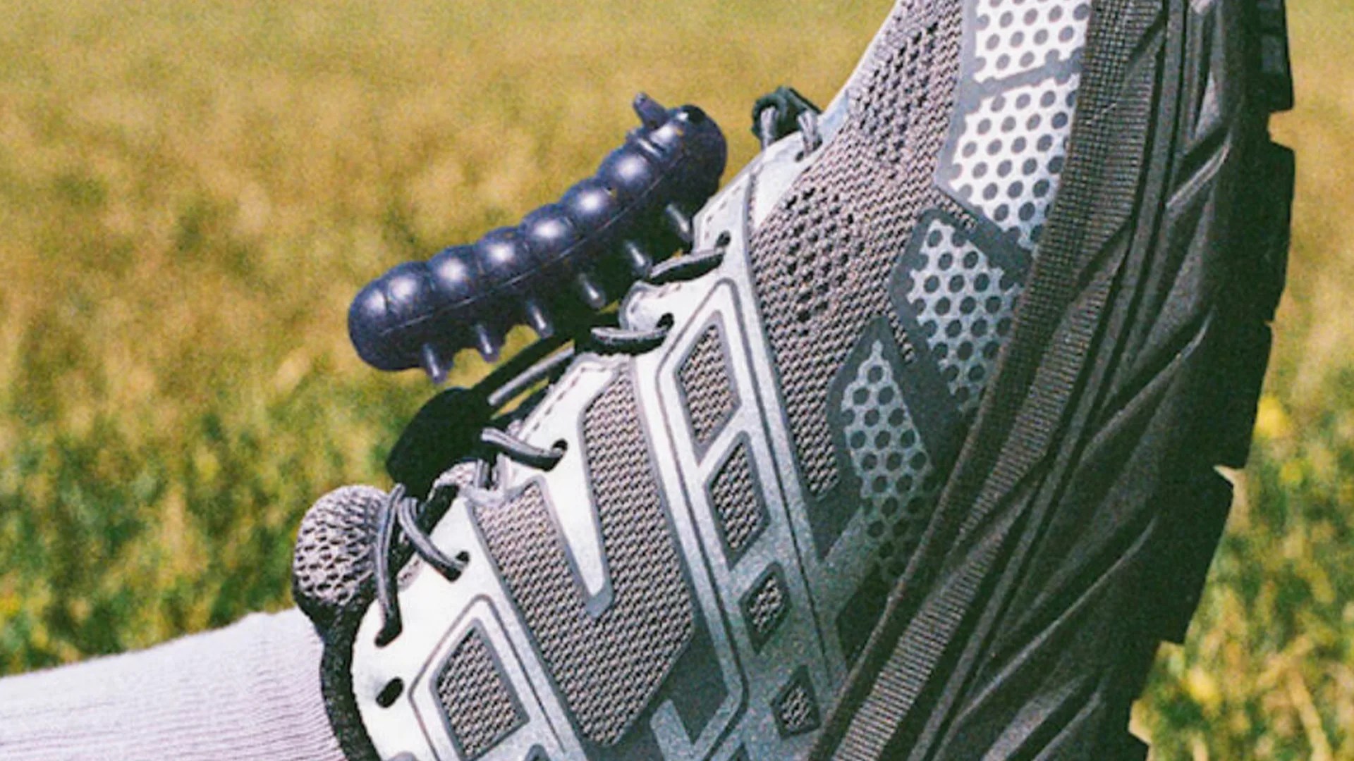 Close-up of a gray and black athletic shoe with a textured mesh design and a thick, rugged sole, resting on grass. A black, segmented massage roller is placed on top of the shoe near the laces.