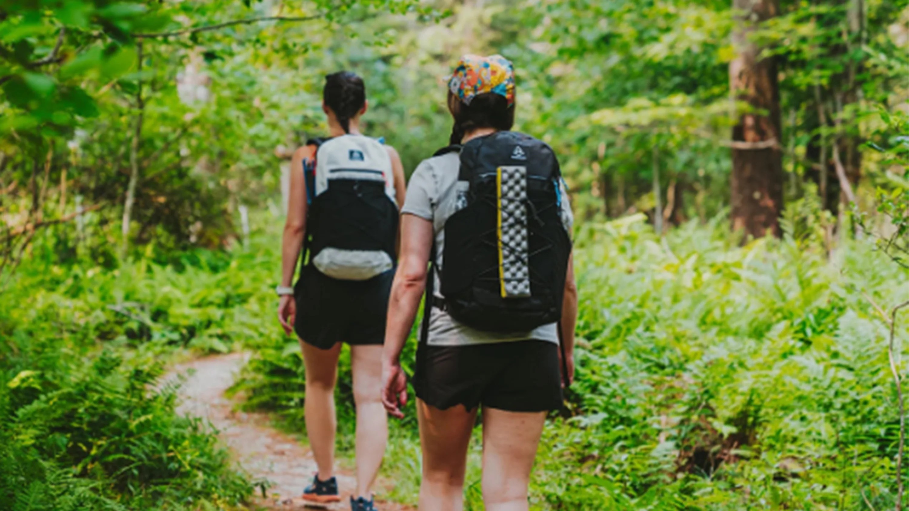 Two people hiking on a narrow dirt trail through a lush green forest, both wearing backpacks and casual outdoor clothing.