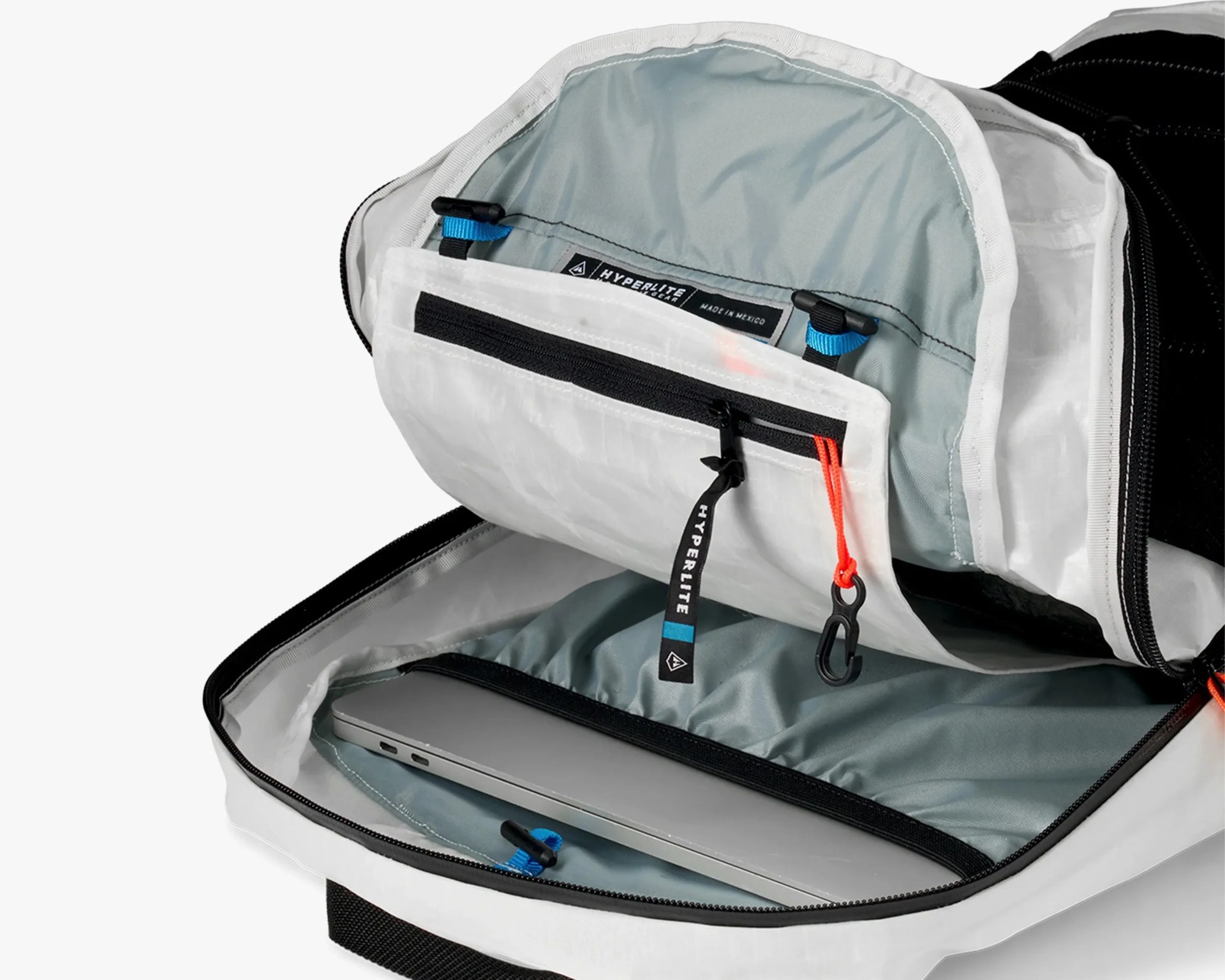 Open white and black backpack showing interior compartments, including a padded laptop sleeve holding a silver laptop, a zippered pocket with a black zipper pull labeled "HYPERLITE," and a small black clip attached to an orange cord inside the main compartment. The interior lining is light gray with blue and black accents.