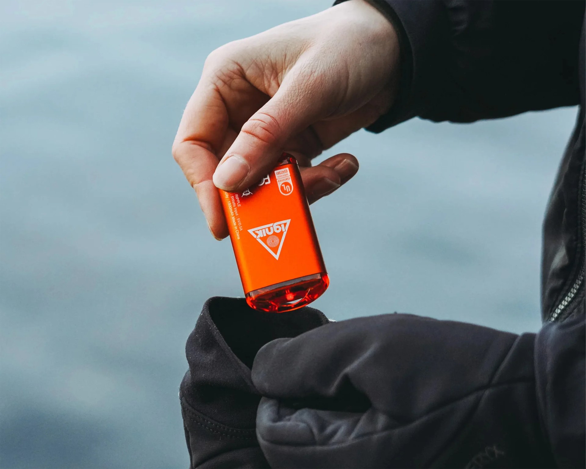 A hand holding a small, bright orange bottle of hand sanitizer above a black glove, with a blurred blue background. The bottle has white text and a triangular logo on it.