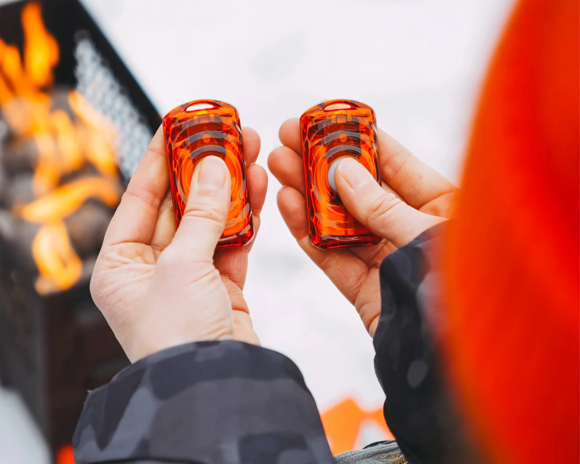 Two hands holding small orange safety flares with a fire burning in the background.