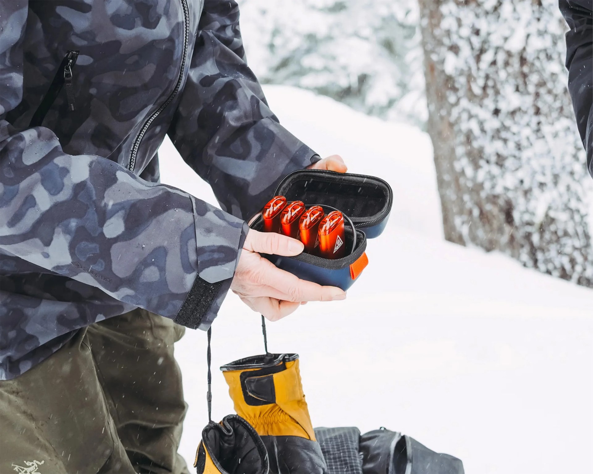A person wearing a dark camouflage jacket and olive green pants is holding an open small black case containing four red-orange devices. The scene is outdoors in a snowy environment, with snow-covered trees in the background. A pair of yellow and black gloves is visible below the case.
