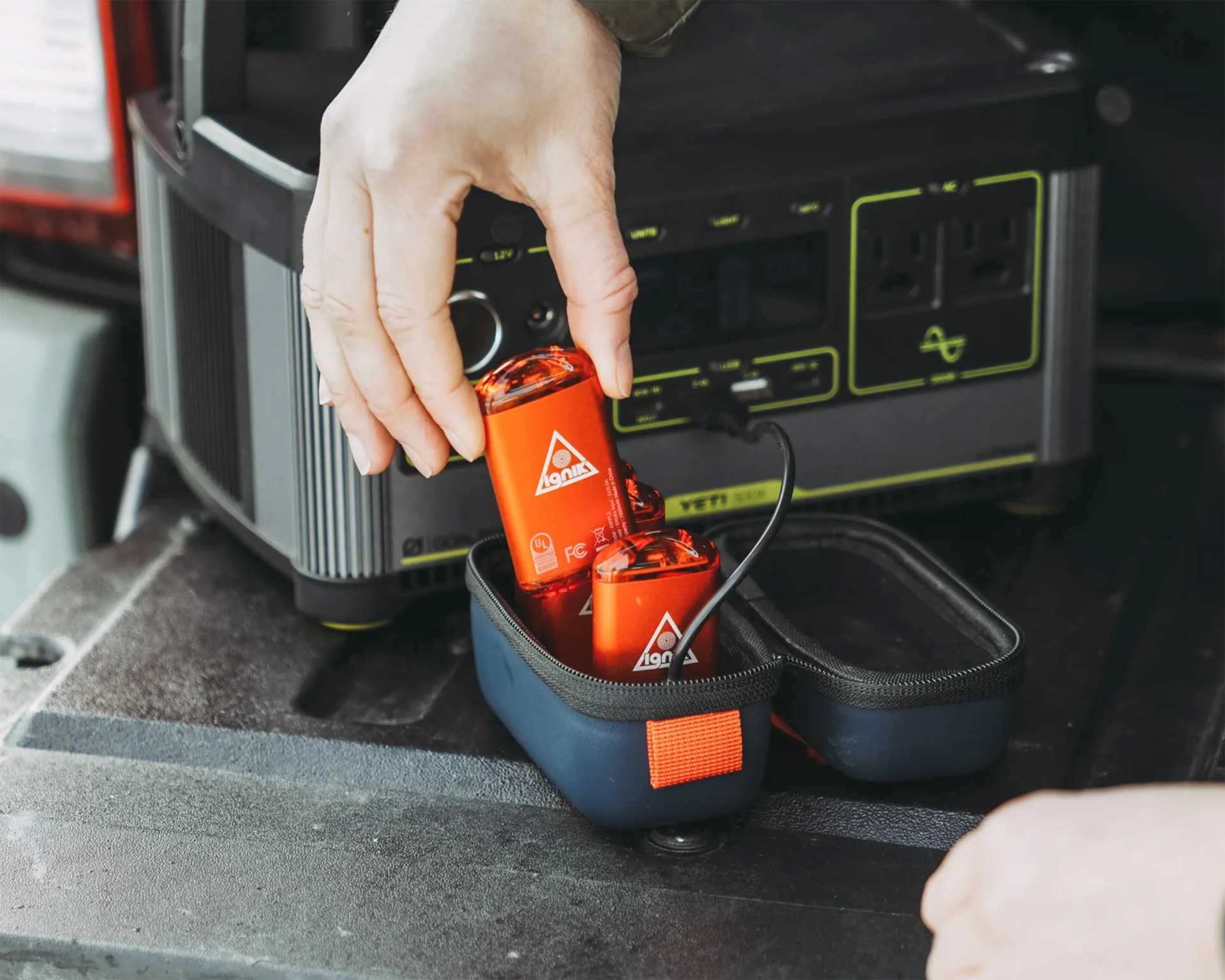 A hand is placing a small red device with a triangular logo into a navy blue zippered case. The case contains similar red devices and is positioned on a dark surface in front of a portable power station with multiple outlets and ports.