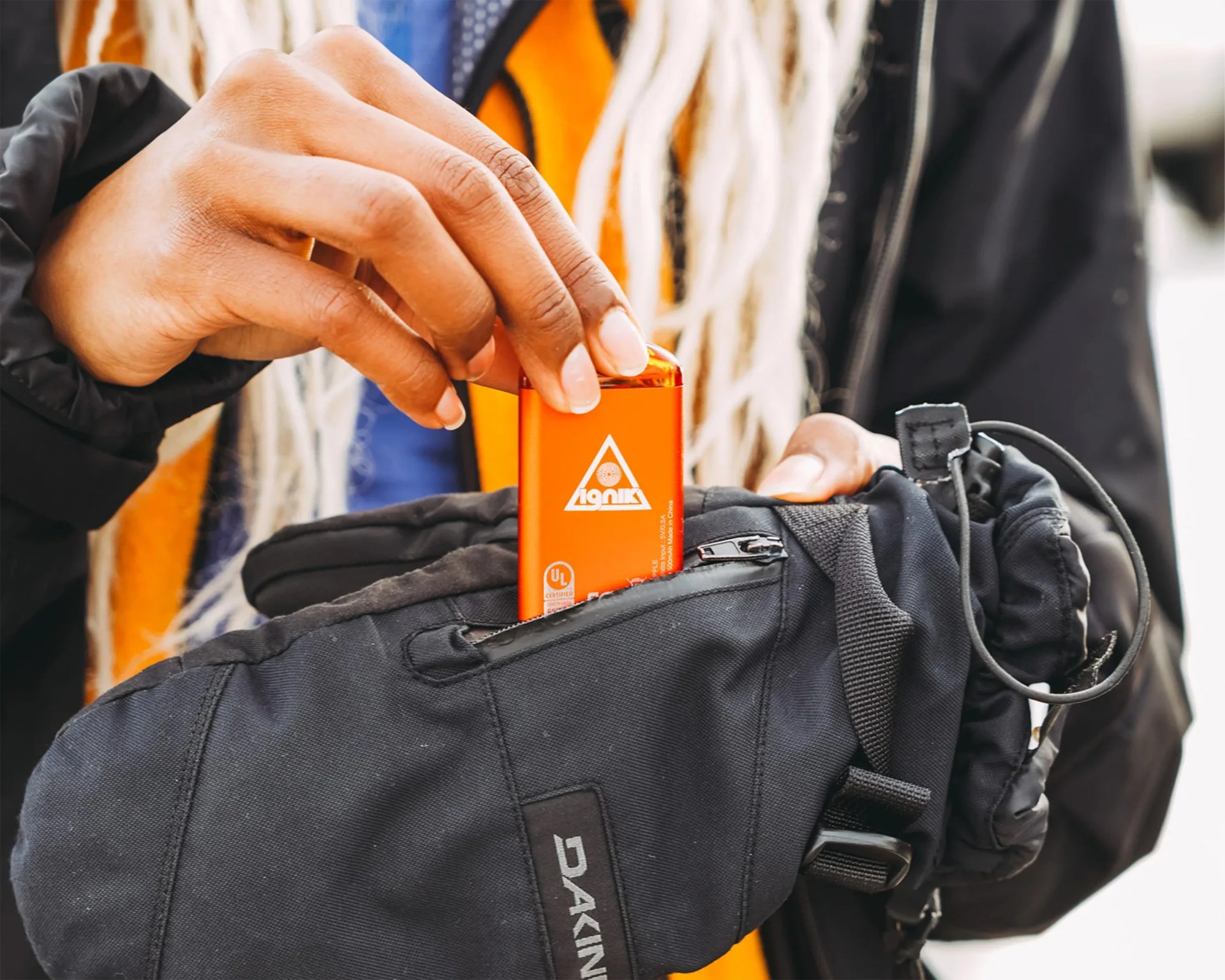 A person is placing an orange rectangular object with a triangular logo and text into the zippered pocket of a black Dakine glove. The person has light brown skin and long, light-colored hair. They are wearing a black jacket over an orange garment.