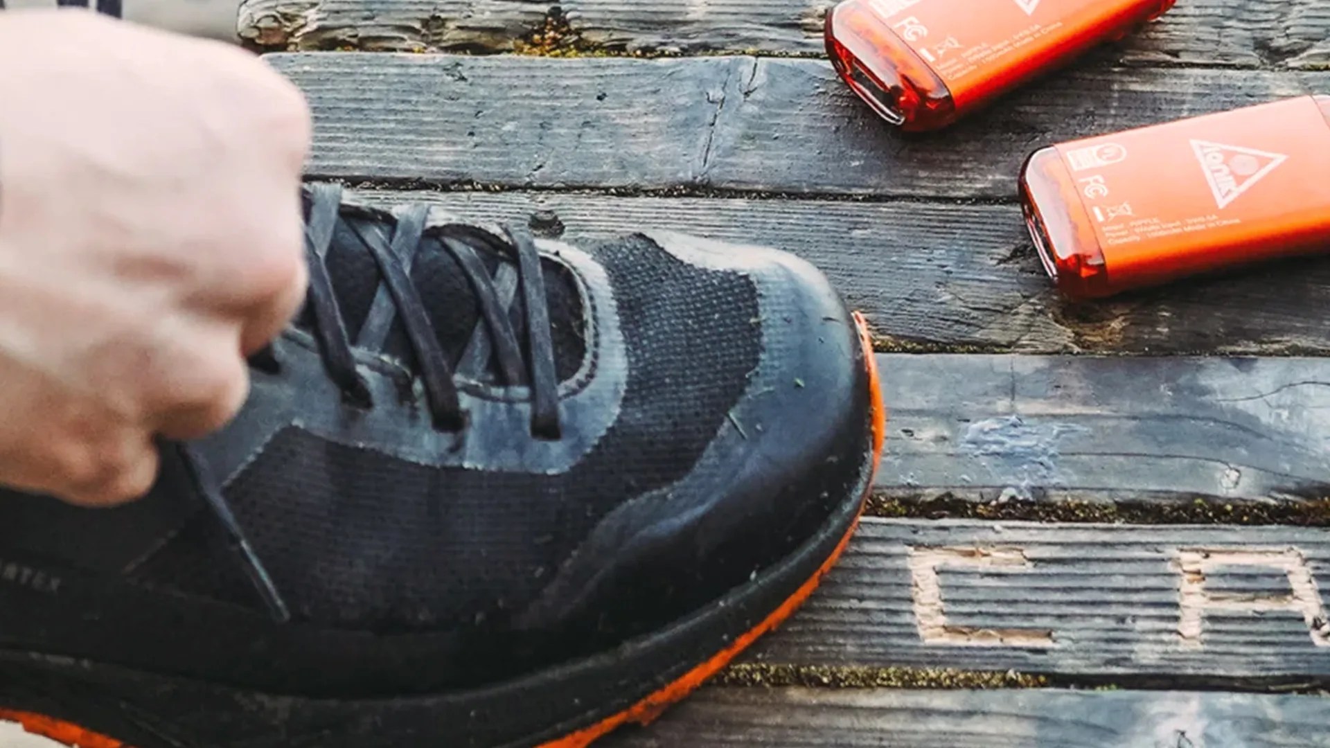 A close-up of a black and orange athletic shoe being tied on a weathered wooden surface. Two orange rectangular objects with white markings are placed nearby on the wood.