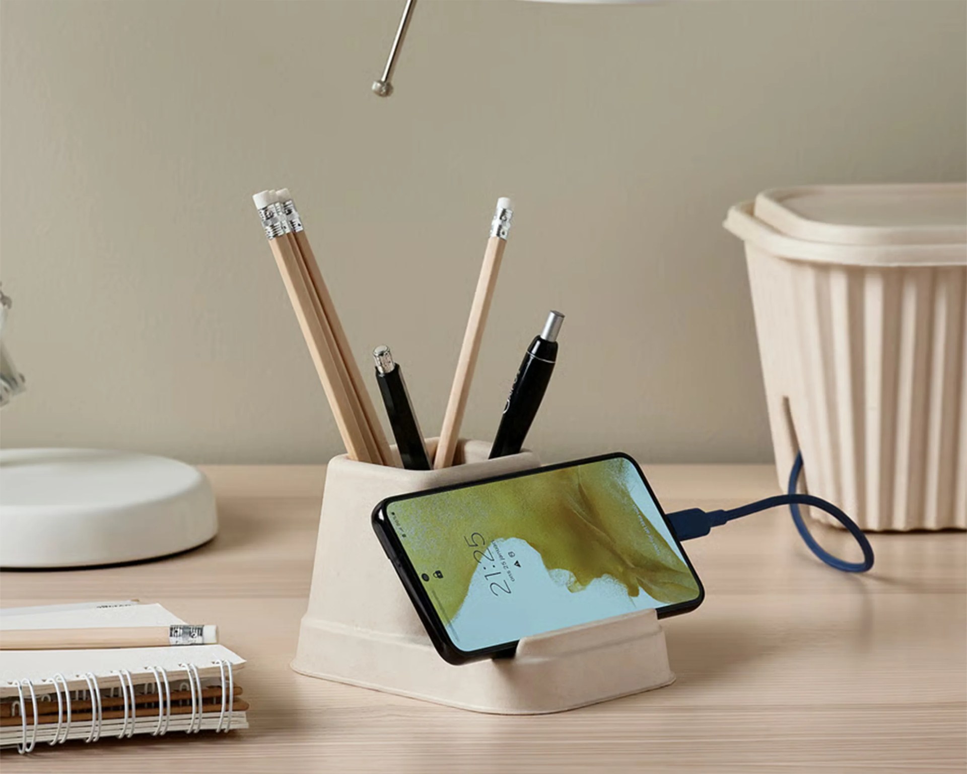 A beige desk organizer holding several pencils and pens, with a smartphone resting horizontally in a slot at the front, connected to a charging cable. The organizer is placed on a light wooden desk, alongside a spiral notebook and a beige container in the background.