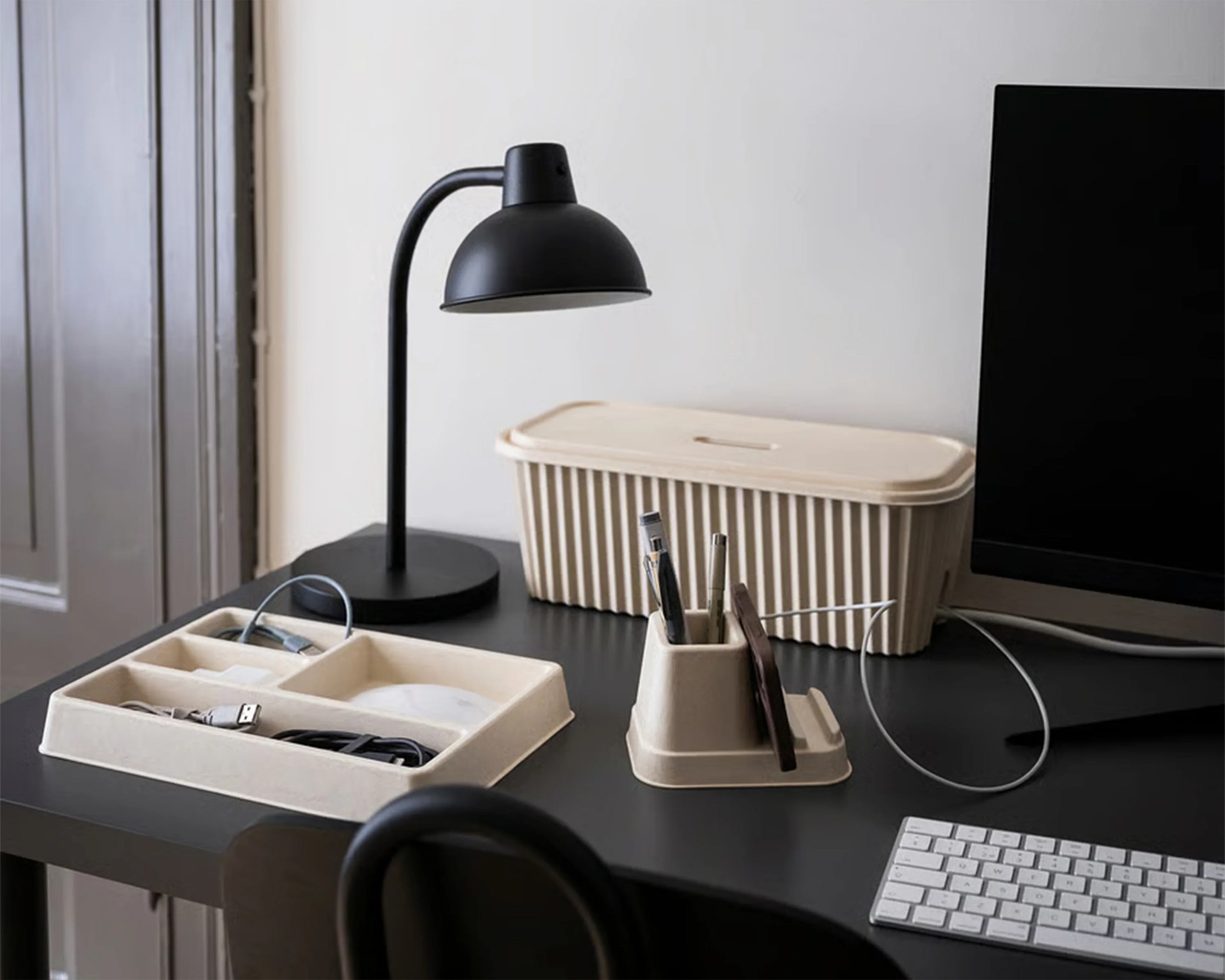 A black desk lamp with a curved neck sits on a black desk next to a beige rectangular storage box with a lid. In front of the storage box is a beige organizer holding pens and a smartphone. To the left, there is a beige tray with compartments containing cables and a USB drive. A white keyboard and part of a computer monitor are also visible on the desk.