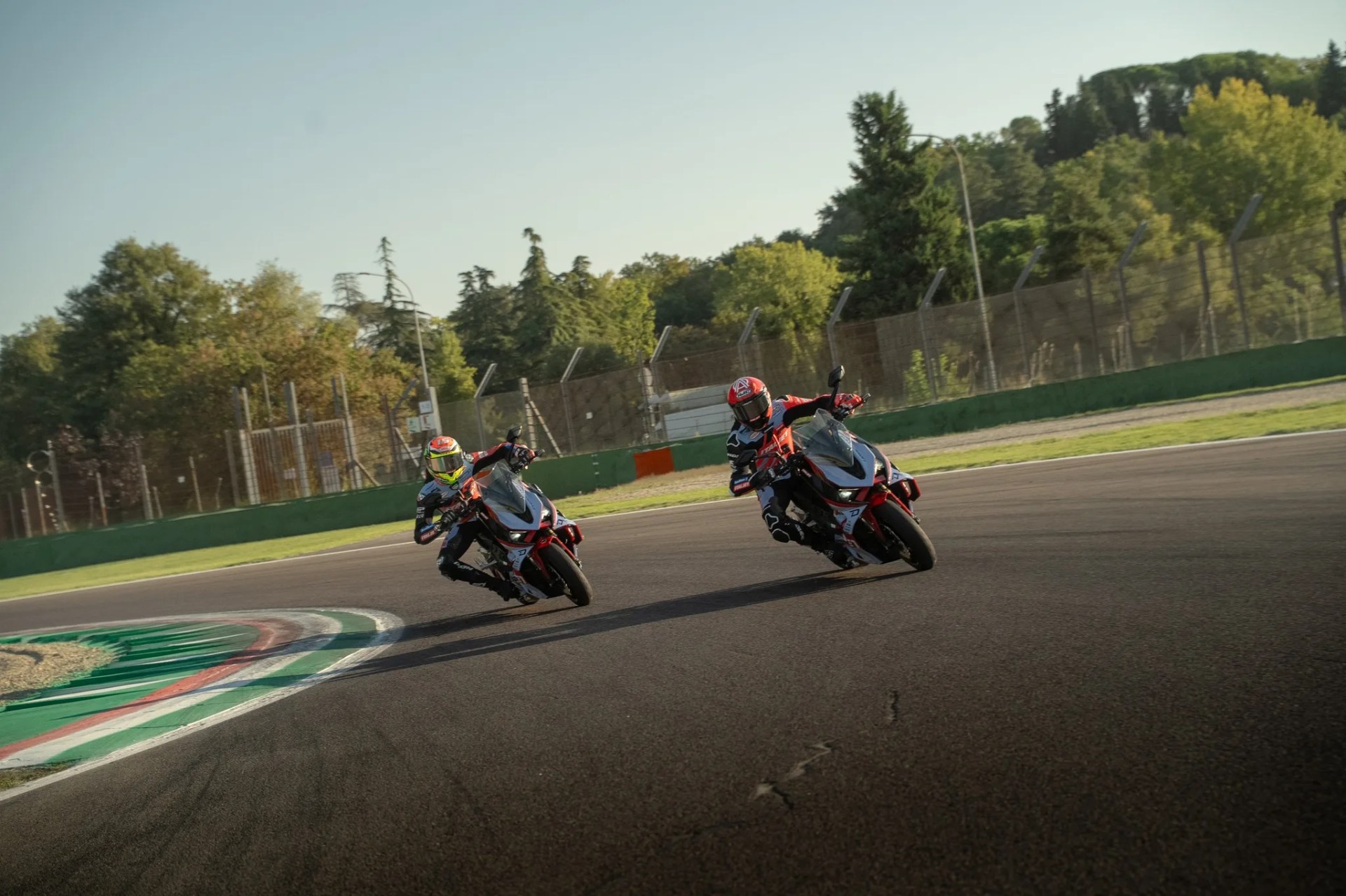 Two motorcyclists wearing helmets and racing suits are leaning into a curve on a racetrack, riding sport motorcycles with red and black accents. The track has green, white, and red curbing on the left side, and there is a fence and trees in the background under a clear sky.