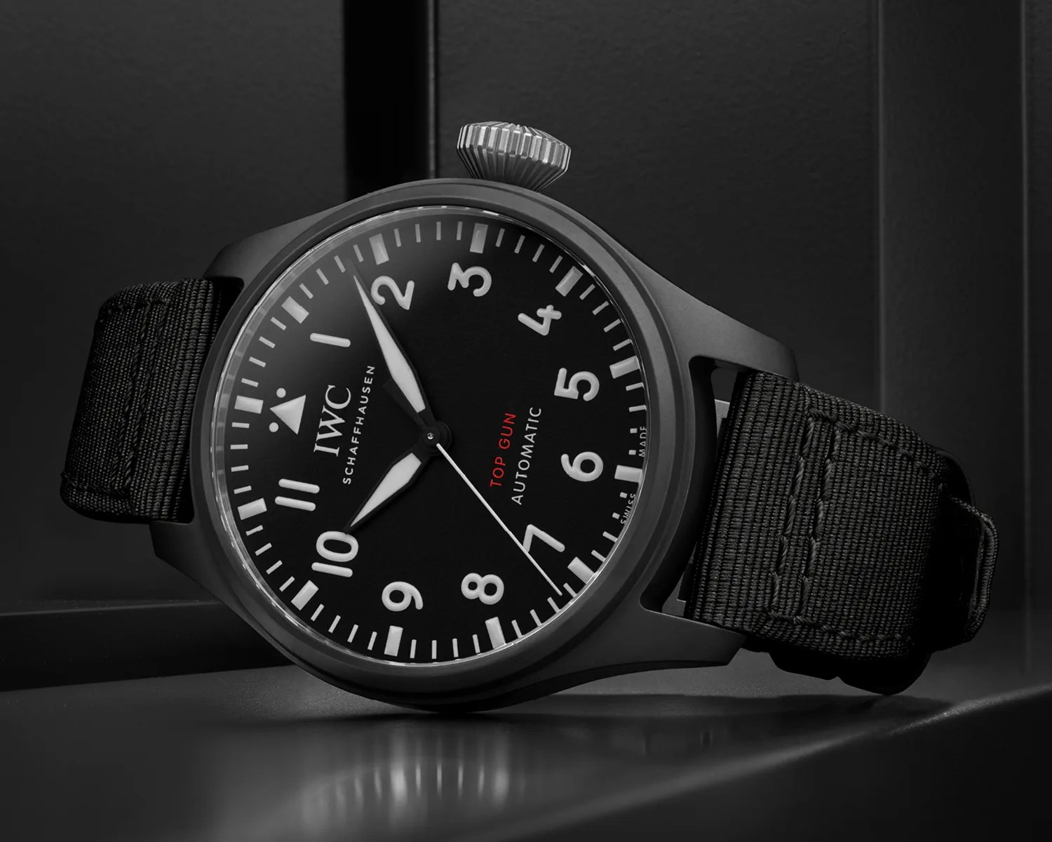 Black IWC Schaffhausen Top Gun Automatic wristwatch with a black fabric strap, white numerals, and hands, and a red "TOP GUN" inscription on the dial. The watch is positioned on a reflective dark surface against a dark background.