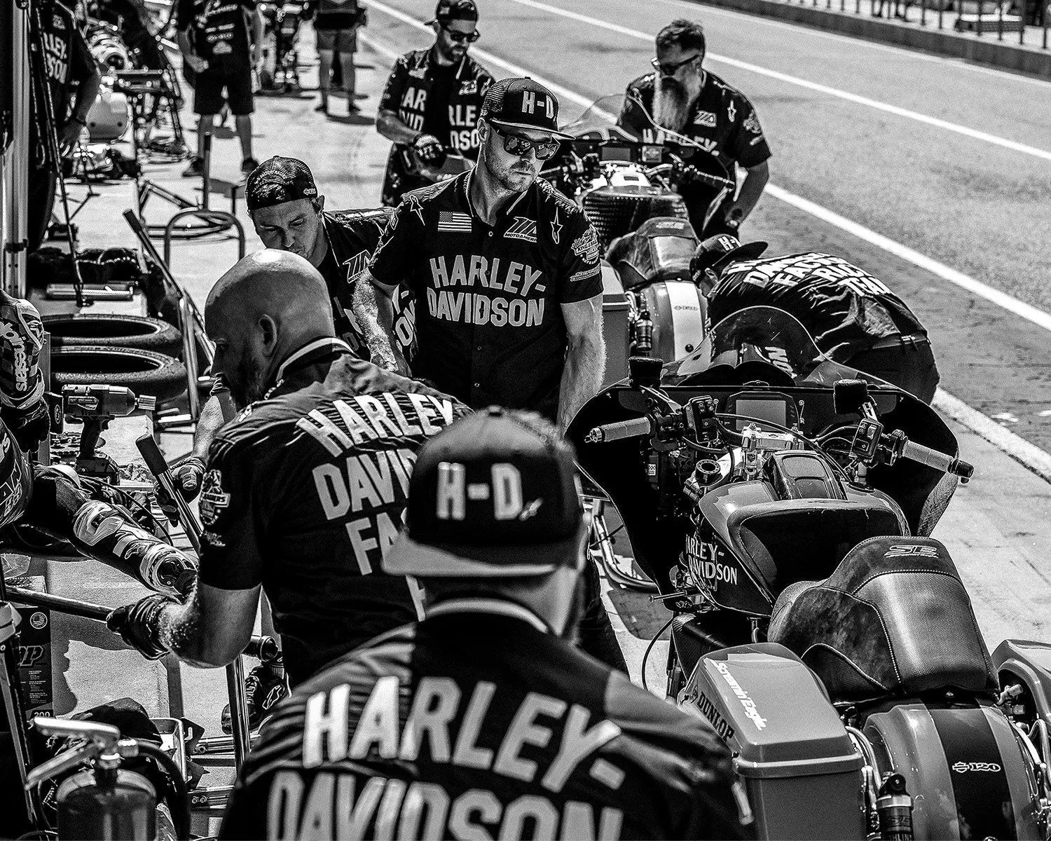 A black and white photo of a Harley-Davidson racing pit crew working on motorcycles. Several crew members wear matching Harley-Davidson shirts and caps, focused on tasks around the bikes. The scene is busy with tools, tires, and equipment visible, set on a race track pit lane.