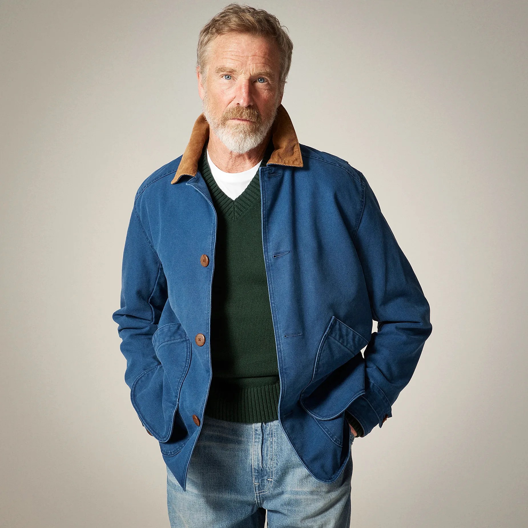A middle-aged man with a beard wearing a blue jacket with a brown corduroy collar, a dark green V-neck sweater over a white shirt, and light blue jeans. The jacket has large front pockets and brown buttons. The man has his hands in the jacket pockets and is standing against a plain light gray background.