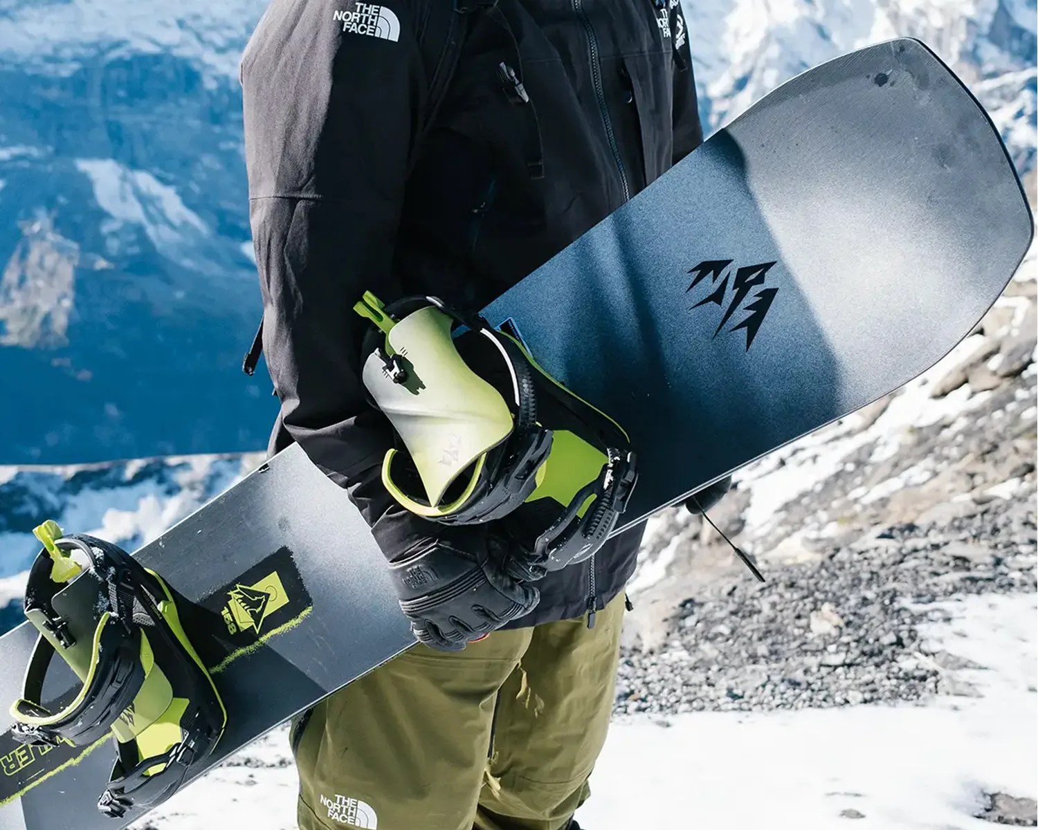 A person wearing a black jacket and olive green pants, both branded with The North Face logo, is holding a black snowboard with neon green bindings. The background shows a snowy mountainous landscape.