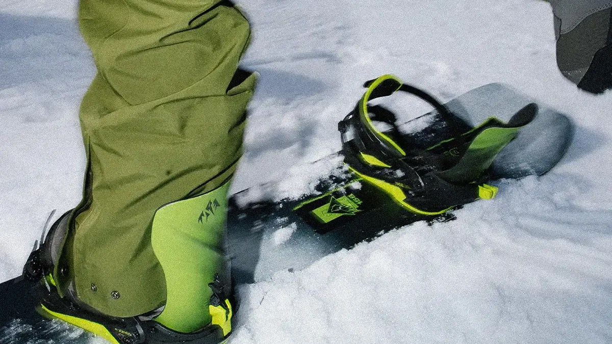 A close-up of a snowboard with black and neon green bindings and boots, partially buried in snow. The snowboard has a black base with neon green accents, and the boots are neon green with black details. The person wearing the boots is dressed in olive green snow pants.