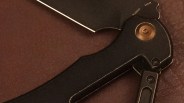 Close-up of a black folding knife handle with a bronze pivot screw and visible screws securing the handle. The blade is partially visible, showing a sharp edge and the words "MASER" and "SWEDEN" near the pivot. The background is a textured brown surface.