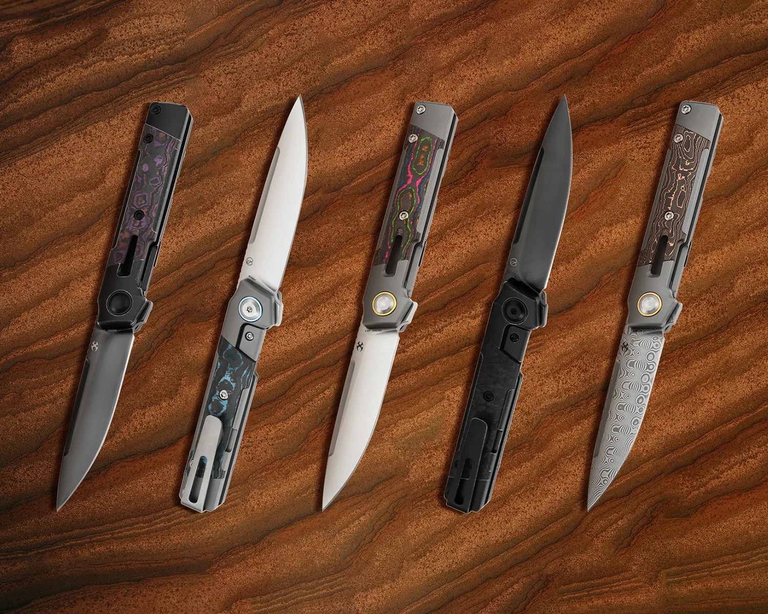 Five folding knives with various blade finishes and handle designs are displayed on a textured brown surface. The knives feature sleek, modern handles with different color accents and patterns, including black, blue, multicolor, and wood-like textures. The blades vary in finish, including polished steel, black-coated, and patterned Damascus steel.