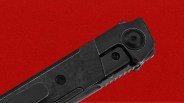 Close-up of the top part of a black folding knife with a textured handle and visible screws, set against a solid red background.