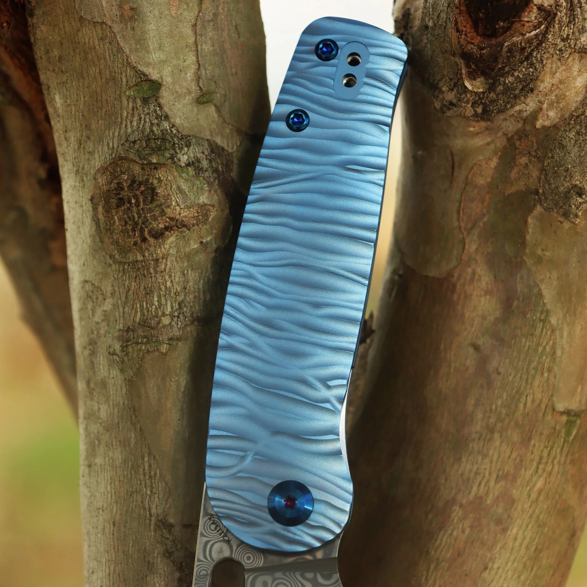 Blue textured handle of a folding knife with visible screws, resting against the bark of a tree. The handle has a wavy, ridged pattern and a metallic finish. Part of the patterned blade is visible at the bottom.