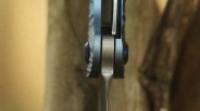 Close-up view of the spine and locking mechanism of a folding knife, showing the textured handle and the blade's back edge. The knife is positioned vertically between two blurred surfaces.
