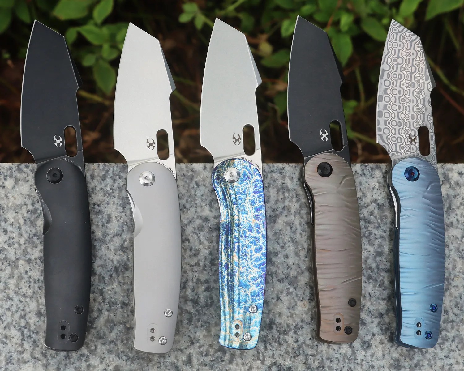 Five folding knives with cleaver-style blades are displayed side by side on a stone surface. The knives have different handle colors and finishes: black, matte silver, blue with a marbled pattern, brown with a textured finish, and light blue with a wavy texture. The blades vary in finish, including black, silver, and a patterned Damascus steel design. Each blade has a small oval thumb hole near the base.