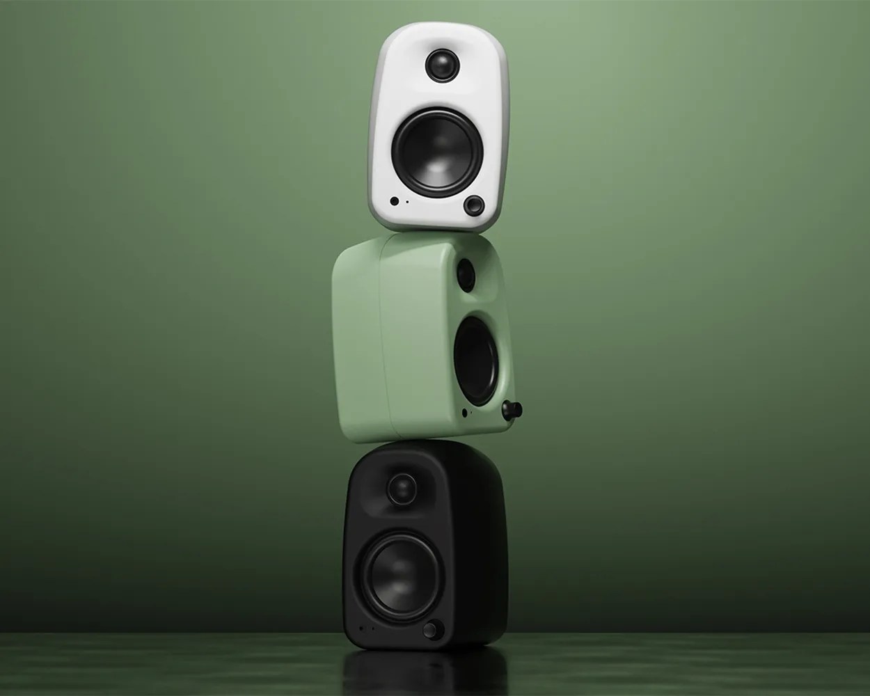 Three compact speakers stacked vertically, with the top speaker in white, the middle speaker in light green, and the bottom speaker in black, all set against a gradient green background. Each speaker features a tweeter and a woofer, with a small control knob on the lower right corner.