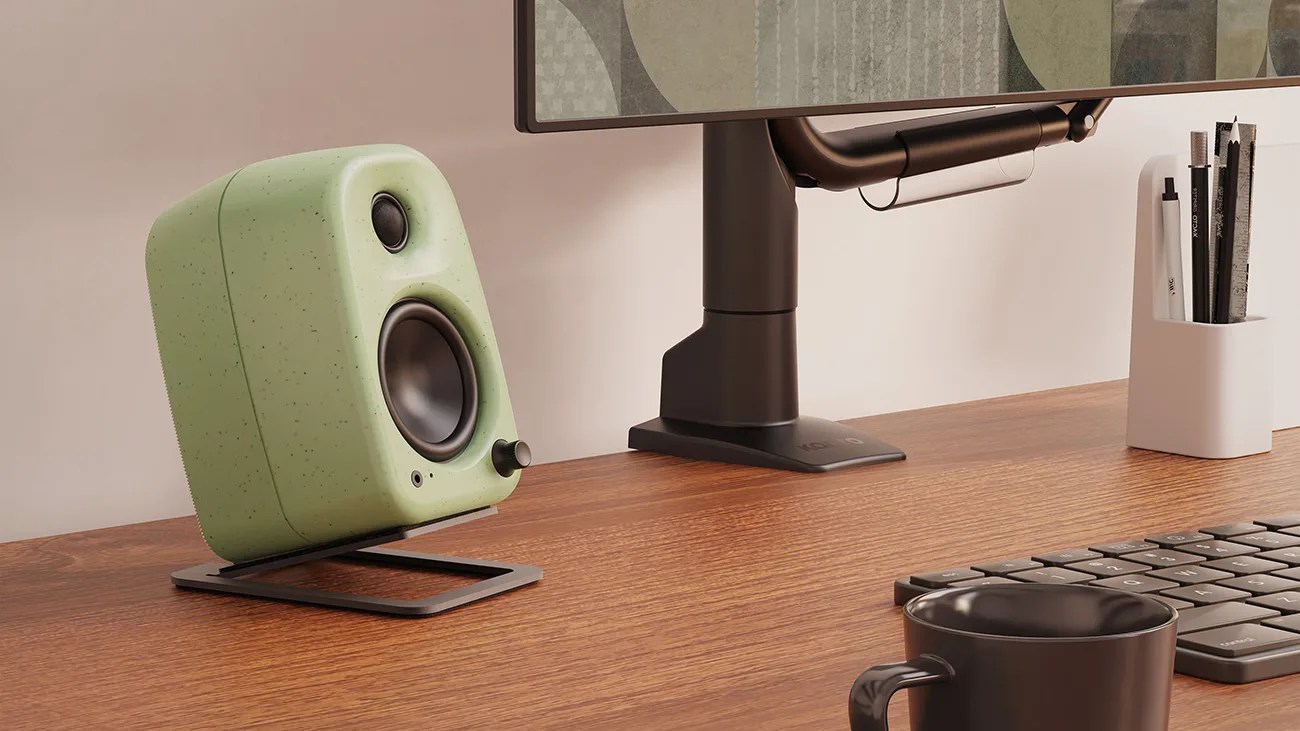 Green desktop speaker with a rounded rectangular shape and black speaker cones, placed on a black angled stand on a wooden desk. Nearby are a black keyboard, a black coffee cup, a white container holding pencils and pens, and the base of a monitor stand.