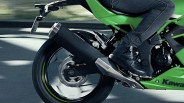 Close-up of the rear section of a green Kawasaki motorcycle in motion, showing the rear wheel, exhaust pipe, and a rider wearing black boots and dark pants. The rear wheel is spinning, creating a blurred effect.