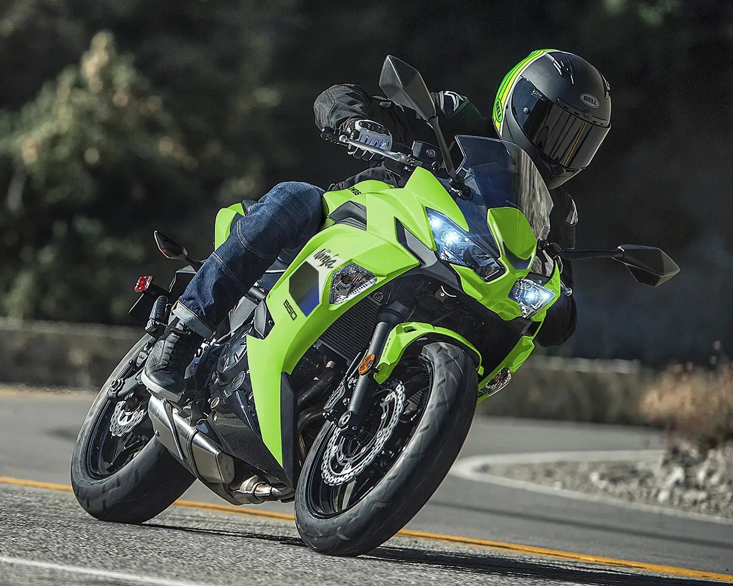 A person wearing a black jacket, blue jeans, black shoes, and a black helmet with green and yellow stripes is riding a bright green Kawasaki Ninja 650 motorcycle, leaning into a turn on a paved road. The motorcycle's headlights are on, and the background is blurred with trees and a stone wall.