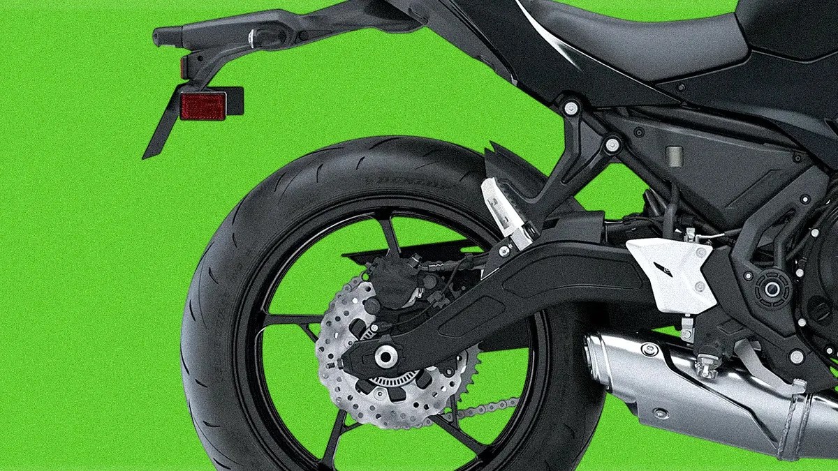 Rear section of a black motorcycle showing the rear wheel with a disc brake, chain drive, exhaust pipe, and part of the seat and frame against a bright green background.