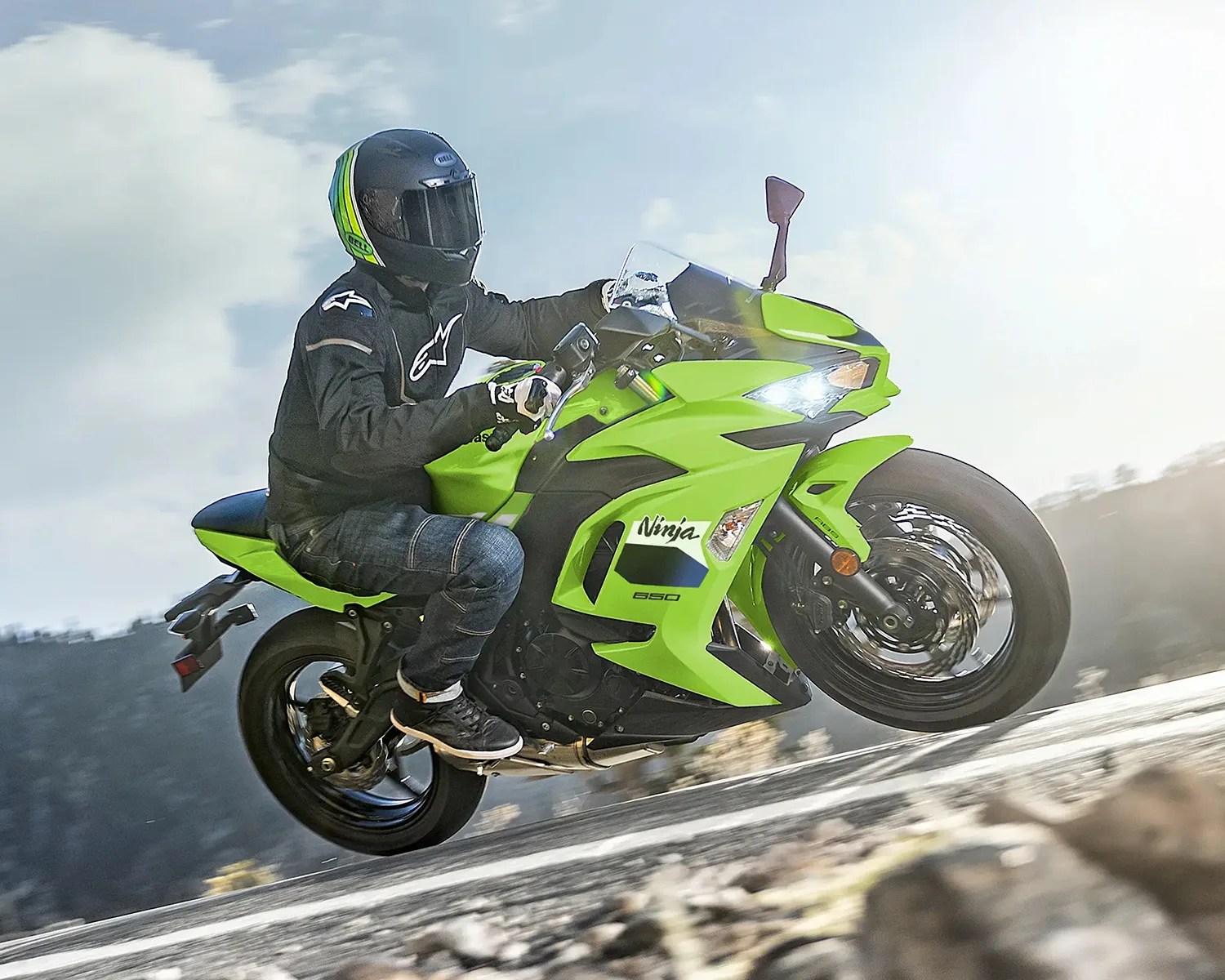 A rider wearing a black helmet with green and yellow stripes, a black jacket, gloves, and jeans is performing a wheelie on a bright green Kawasaki Ninja 650 motorcycle on a paved road with a blurred natural background. The motorcycle's front wheel is lifted off the ground, and the headlight is on.