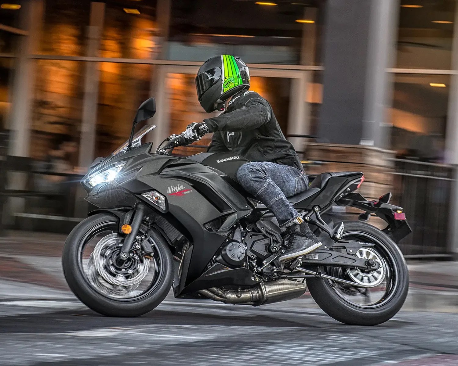 A person wearing a black jacket, blue jeans, black boots, and a black helmet with green stripes is riding a black Kawasaki Ninja 650 motorcycle on a city street at night. The motorcycle's front headlight is on, and the background shows blurred urban lights and buildings.